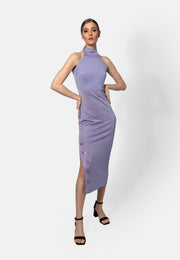 Model trägt ein lilacfarbenes, ärmelloses Midikleid aus glänzendem Viskose-Jersey mit hohem Kragen und Schlitz auf der Seite, verziert mit kleinen roten Stickereien. Das Kleid reicht bis unter das Knie und wird mit schwarzen Sandaletten kombiniert.