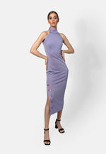 Model trägt ein lilacfarbenes, ärmelloses Midikleid aus glänzendem Viskose-Jersey mit hohem Kragen und Schlitz auf der Seite, verziert mit kleinen roten Stickereien. Das Kleid reicht bis unter das Knie und wird mit schwarzen Sandaletten kombiniert.