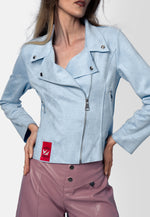 Model trägt eine kurz geschnittene Bikerjacke aus veganem Wildleder in Eisblau mit asymmetrischem Reißverschluss und silbernen Details. Die Jacke wird kombiniert mit einer hochgeschnittenen, fliederfarbenen Hose.
